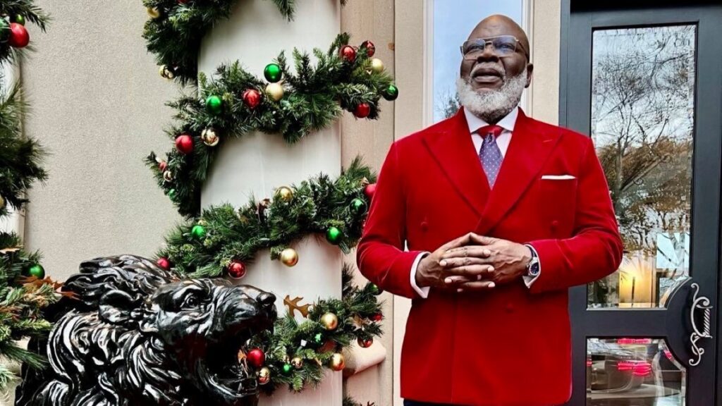 T.D Jakes wearing a red suite standing in the side of a Christmas tree.